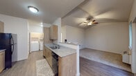 This single-floor unit showcases a thoughtfully designed open-concept layout that connects the kitchen, dining, and living areas into one cohesive space. The kitchen is well-appointed with light wood cabinetry, granite-style countertops, a peninsula with a double sink, and black appliances including an electric range, microwave, and refrigerator. Wood-look plank flooring runs throughout the kitchen and main living areas, complementing the warm cabinetry tones. A vaulted ceiling fitted with a ceiling fan crowns the living area, adding vertical dimension and promoting air circulation. Natural light enters through multiple windows fitted with blinds, including a glass-paneled exterior door that opens to the outdoors. A dedicated in-unit laundry area with a full-size washer and dryer is conveniently tucked off the kitchen. The bedrooms are carpeted and receive natural light through exterior-facing windows. Two bathrooms are present, each featuring granite-topped wood vanities with undermount sinks, tile-style flooring, and clean white walls. One bathroom includes a tub-shower combination, while the other offers a stand-alone shower. The overall aesthetic is modern, neutral, and move-in ready.