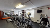 This fitness center is organized across a single level, offering a comprehensive layout divided into distinct workout zones. The cardio area is lined with multiple treadmills and elliptical cross-trainers, all equipped with individual digital consoles for tracking workout metrics. Rubber flooring throughout the space provides durability and shock absorption underfoot. Natural light filters in through several windows fitted with blinds, complementing the overhead fluorescent panel lighting that keeps the space well-illuminated.

A large wall mirror spans a significant portion of the main workout area, visually expanding the space and allowing users to monitor their form during exercise. A wall-mounted television is positioned for viewing during cardio sessions. The strength training section is set apart by its bold orange accent walls, creating a visually distinct zone. This area houses a multi-station cable machine, a weight rack, and a flat bench, providing options for a variety of resistance exercises. A vending machine is also present, offering convenience for users. The textured ceiling and clean, practical aesthetic throughout give the fitness center a functional and straightforward character suited for daily use.