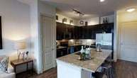 This single-floor space showcases a thoughtfully designed interior centered around a striking modern kitchen. The kitchen is a standout feature, boasting rich dark espresso cabinetry that extends to the upper walls, creating a bold contrast against the light-toned walls and granite countertops with warm, mottled tones. A peninsula with a breakfast bar overhang provides additional seating and prep space, making it ideal for both cooking and casual dining. Stainless steel appliances, including a refrigerator with an ice and water dispenser, a microwave, and a range with an electric cooktop, lend a contemporary and polished feel. A classic tile backsplash adds texture and detail to the cooking area, while track lighting overhead ensures the workspace is well-lit. A full-panel pantry door offers practical storage discreetly tucked beside the cabinetry. Rich dark hardwood-style flooring runs throughout, grounding the space with warmth and cohesion. Decorative accents atop the upper cabinets and along the counters add a curated, homey touch. The overall ambiance is sleek and modern, with a balance of dark and light tones creating an inviting and functional living environment.