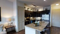 This single-floor space showcases a thoughtfully designed interior centered around a striking modern kitchen. The kitchen is a standout feature, boasting rich dark espresso cabinetry that extends to the upper walls, creating a bold contrast against the light-toned walls and granite countertops with warm, mottled tones. A peninsula with a breakfast bar overhang provides additional seating and prep space, making it ideal for both cooking and casual dining. Stainless steel appliances, including a refrigerator with an ice and water dispenser, a microwave, and a range with an electric cooktop, lend a contemporary and polished feel. A classic tile backsplash adds texture and detail to the cooking area, while track lighting overhead ensures the workspace is well-lit. A full-panel pantry door offers practical storage discreetly tucked beside the cabinetry. Rich dark hardwood-style flooring runs throughout, grounding the space with warmth and cohesion. Decorative accents atop the upper cabinets and along the counters add a curated, homey touch. The overall ambiance is sleek and modern, with a balance of dark and light tones creating an inviting and functional living environment.