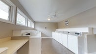 Located at 18 Tall Pine Dr in Hampton, Virginia, this single-floor laundry facility is designed to accommodate communal use with an efficient, functional layout. The room is lined on both sides with rows of white washing machines and dryers, providing a generous number of units to serve multiple users simultaneously. A folding counter is positioned near the entrance, offering a convenient surface for handling laundry. The space features a speckled, easy-to-clean floor, which adds durability suited to high-traffic use. Natural light filters in through a row of horizontal windows along one wall, creating a bright and open atmosphere. A ceiling fan mounted at the center of the sloped white ceiling promotes air circulation throughout the room. The walls are finished in a neutral beige tone, contributing to a clean and straightforward aesthetic. Informational postings are displayed on the wall, providing users with relevant guidelines. Overall, the facility is well-organized, well-lit, and equipped to meet the everyday laundry needs of residents.
