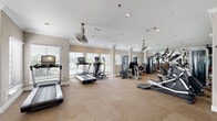 This fitness center is a thoughtfully designed workout facility that offers a full range of exercise options within a spacious, single-floor layout. The space is divided into distinct functional zones, allowing for seamless transitions between cardio training, strength work, and stretching.

The cardio section is lined with multiple treadmills, elliptical machines, and a recumbent bike, all positioned along large windows fitted with horizontal blinds. These windows flood the area with natural light and provide views of an outdoor pool area, creating an energizing atmosphere during workouts. Ceiling fans with industrial-style finishes are suspended overhead, keeping the space well-ventilated.

The strength training zone features a robust selection of cable machines, multi-function weight stations, and selectorized equipment arranged across a carpeted floor. Full-length mirrors line the walls of this section, visually expanding the space and allowing users to monitor their form. A dedicated free weights area includes a rack of dumbbells in various sizes, a flat utility bench, and wall-mounted yoga mats for stretching.

Throughout the facility, neutral warm-toned walls, crown molding, and recessed lighting contribute to a clean, polished aesthetic. A colorful rack of stability balls adds a lively accent near one corner. The overall ambiance is bright, functional, and welcoming, making it a well-rounded fitness amenity.