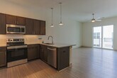 Located at 590 Downey Green St in Hampton, Virginia, this single-floor unit presents a clean, contemporary design throughout. Light wood-tone flooring runs continuously across the living spaces, lending a cohesive and airy feel to the interior. The open-concept layout connects the main living area to the kitchen, maximizing the sense of space and encouraging a functional flow between rooms.

The kitchen is well-appointed with rich dark wood cabinetry, stainless steel appliances including a refrigerator, range, and microwave, and a granite-topped peninsula with pendant lighting overhead. A tiled backsplash adds a refined finishing touch to the cooking space. In-unit laundry is conveniently tucked behind panel doors nearby.

The living area benefits from natural light streaming through a sliding glass door that opens to a private balcony with exterior views and railing. Ceiling fans are installed throughout the main rooms, providing both comfort and additional lighting. The bedroom features a large window overlooking the exterior and direct access to a walk-in closet. The bathroom is straightforward and functional, offering a soakng tub with shower, wood-tone flooring, and an adjacent walk-in closet. White walls and clean lines define the overall aesthetic of this modern unit.