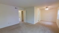 This single-floor residential unit offers a clean, functional layout designed for comfortable everyday living. The main living area is finished with soft, neutral-toned wall-to-wall carpeting that extends throughout the space, creating a warm and cohesive feel underfoot. White walls and ceiling surfaces contribute to a bright, airy ambiance, while a ceiling fan provides both lighting and ventilation to the space.

The layout opens naturally toward multiple adjoining rooms, with clearly defined doorways leading to additional areas of the unit. A spacious closet with visible shelving and hanging rod is accessible from the main living area, offering ample built-in storage. Overhead HVAC vents are integrated into the ceiling, ensuring climate comfort year-round.

A glimpse into the adjacent area reveals a full-size refrigerator, indicating a kitchen or kitchenette nearby. The open sightlines between rooms lend the space a sense of connectivity and flow, making it feel larger than its footprint suggests. The overall aesthetic is neutral and move-in ready, with a simple, understated design that provides a versatile canvas for personalization. This well-maintained unit balances practical storage solutions with comfortable, light-filled living spaces.