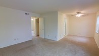 This single-floor residential unit offers a clean, functional layout designed for comfortable everyday living. The main living area is finished with soft, neutral-toned wall-to-wall carpeting that extends throughout the space, creating a warm and cohesive feel underfoot. White walls and ceiling surfaces contribute to a bright, airy ambiance, while a ceiling fan provides both lighting and ventilation to the space.

The layout opens naturally toward multiple adjoining rooms, with clearly defined doorways leading to additional areas of the unit. A spacious closet with visible shelving and hanging rod is accessible from the main living area, offering ample built-in storage. Overhead HVAC vents are integrated into the ceiling, ensuring climate comfort year-round.

A glimpse into the adjacent area reveals a full-size refrigerator, indicating a kitchen or kitchenette nearby. The open sightlines between rooms lend the space a sense of connectivity and flow, making it feel larger than its footprint suggests. The overall aesthetic is neutral and move-in ready, with a simple, understated design that provides a versatile canvas for personalization. This well-maintained unit balances practical storage solutions with comfortable, light-filled living spaces.