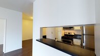 This single-floor space offers a clean and functional layout designed for comfortable everyday living. The kitchen is a standout feature, showcasing a pass-through design with a dark, reflective countertop that serves as both a workspace and casual bar. Stainless steel appliances — including a gas range and over-the-range microwave — lend a modern, polished look, while light cabinetry provides ample storage. The pass-through opening connects the kitchen visually to the rest of the space, creating an open and airy feel that encourages easy interaction between rooms. Rich hardwood flooring runs throughout, adding warmth and continuity to the interior. The walls are finished in crisp white, allowing natural light to reflect and brighten the space. A hallway leads from the main living area to additional rooms, suggesting a well-organized floor plan. The entry door, visible through the kitchen pass-through, reinforces the compact yet efficient use of space. Overall, the interior strikes a balance between contemporary design and practical functionality, making it an inviting and well-appointed living environment.