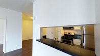 This single-floor space offers a clean and functional layout designed for comfortable everyday living. The kitchen is a standout feature, showcasing a pass-through design with a dark, reflective countertop that serves as both a workspace and casual bar. Stainless steel appliances — including a gas range and over-the-range microwave — lend a modern, polished look, while light cabinetry provides ample storage. The pass-through opening connects the kitchen visually to the rest of the space, creating an open and airy feel that encourages easy interaction between rooms. Rich hardwood flooring runs throughout, adding warmth and continuity to the interior. The walls are finished in crisp white, allowing natural light to reflect and brighten the space. A hallway leads from the main living area to additional rooms, suggesting a well-organized floor plan. The entry door, visible through the kitchen pass-through, reinforces the compact yet efficient use of space. Overall, the interior strikes a balance between contemporary design and practical functionality, making it an inviting and well-appointed living environment.