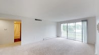 This single-floor residence offers three bedrooms and two bathrooms arranged in a clean, functional layout with a consistently neutral palette throughout. The living area is generously sized, featuring plush carpeting, a sliding glass door that opens to a balcony with outdoor views of surrounding greenery, and a ceiling ventilation system for climate control. Baseboard heating runs along the perimeter, a detail echoed in every carpeted room.

The kitchen stands out with white shaker-style cabinetry, dark granite countertops, wood-look vinyl plank flooring, and black appliances including a dishwasher, electric range, and over-the-range microwave. The layout provides ample counter and storage space.

Each of the three bedrooms is carpeted and brightened by natural light from windows with blinds. Baseboard heating is present throughout, and one bedroom also features a wall-mounted air conditioning unit. The rooms offer comfortable proportions well-suited for various furniture arrangements.

The two bathrooms are finished in white, each featuring a tub-shower combination with a curtain, a toilet, vanity with mirror, and overhead lighting. One bathroom includes a floating-style vanity, adding a contemporary touch to the otherwise classic design.