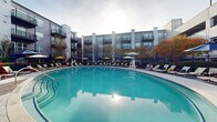 Located at 113 Ellington Blvd in Gaithersburg, Maryland, this outdoor amenity space centers around a generously sized oval swimming pool with striking turquoise water. The pool's smooth, curved design creates an inviting focal point, while the surrounding deck offers an abundance of lounge chairs arranged to maximize sun exposure and relaxation. Colorful umbrellas in shades of navy blue and bright yellow are positioned throughout the deck, providing ample shade and a vibrant visual contrast against the neutral pavement. An ADA-compliant pool lift is conveniently located at the pool's edge, ensuring accessibility for all users. The space is enclosed by a secure perimeter fence, lending a sense of privacy and safety. Lush seasonal trees and manicured landscaping line the borders of the area, softening the transition between the deck and the surrounding multi-story residential buildings, whose balconies overlook the pool courtyard. The open-sky setting allows for generous natural light throughout the day, creating a bright and welcoming atmosphere. The thoughtful layout of seating, shade structures, and greenery makes this outdoor pool area a well-appointed communal retreat suited for both leisurely lounging and active recreation.