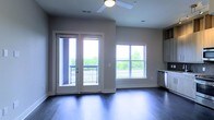 This single-floor space showcases a clean, contemporary design with an open layout that feels airy and inviting. The living area is anchored by rich, dark hardwood flooring that contrasts elegantly with the neutral gray walls, creating a polished, modern aesthetic throughout.

A standout feature is the set of white-trimmed French doors that open directly onto a private balcony, offering outdoor views of lush greenery. The doors are flanked by a large multi-pane window, ensuring the interior is bathed in abundant natural light, giving the space a bright and open atmosphere.

The ceiling is fitted with a ceiling fan, contributing to both comfort and airflow. Recessed vents are integrated seamlessly into the ceiling, maintaining the clean lines of the design. The trim work throughout is crisp and white, providing a sharp contrast to the warm tones of the flooring.

Along one wall, cabinetry is partially visible, suggesting a kitchen or utility area nearby, hinting at a functional and well-organized layout. Overall, the space offers a well-lit, modern environment with thoughtful architectural details and a seamless connection to the outdoors.