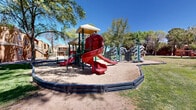This outdoor community space at 9300 Montana Ave in El Paso, Texas, offers a well-appointed recreational area designed to serve residents of the surrounding multi-building complex. The grounds are generously landscaped with mature trees that provide ample shade across the expansive open lawn, creating a welcoming environment suited to a variety of outdoor activities.

A standout feature of the space is the fully equipped playground area, complete with a multi-level play structure featuring vibrant red spiral and straight slides, climbing elements, and decorative rock-style formations. The playground is set within a defined sand boundary bordered by rubber edging, offering a safe and engaging environment for children. In-ground trampolines are also present nearby, adding to the recreational offerings.

A covered picnic pavilion with a tiled roof anchors another area of the grounds, providing shaded seating with picnic tables and a grill — ideal for outdoor gatherings. The surrounding lawn is well-maintained and broad, offering open space between the pavilion, playground, and the warm-toned, multi-story residential buildings that frame the perimeter. Walkways and landscaped borders connect the various areas of the grounds cohesively. Under the clear blue El Paso sky, this community outdoor space presents a functional and inviting environment for recreation and social activity.