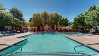 This two-story residential community, branded as Union, offers a thoughtfully designed outdoor amenity space centered around a generously sized swimming pool with crystal-clear turquoise water. The pool area features clearly marked depth indicators and is surrounded by a wide concrete deck furnished with blue lounge chairs, providing ample space for sunbathing and relaxation. A dedicated clubhouse building anchors one end of the pool area, featuring a clean, white stucco exterior with a distinctive arched entrance bearing the Union logo, along with restroom and facility access.

The grounds are richly landscaped with mature shade trees that cast dappled light across the walkways, creating a cool and inviting outdoor atmosphere. Decomposed granite and native low-water plantings are integrated throughout the landscape, reflecting a desert-conscious design aesthetic. Paved walkways wind through the property, connecting the pool area to surrounding garden seating spaces outfitted with blue patio chairs and tables. A wrought iron fence lines portions of the perimeter, adding a sense of enclosure and security. The two-story residential buildings that frame the courtyard feature a neutral stucco facade with exterior staircases, consistent with a Southwestern architectural style. The overall ambiance is open, airy, and well-maintained.