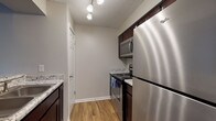 This single-floor residence offers a well-organized layout with distinct living, sleeping, kitchen, and bathroom areas. The kitchen is efficiently designed with dark cabinetry, granite-style countertops, a stainless steel refrigerator, electric range, and over-the-range microwave. Track lighting illuminates the space, and a double-basin sink sits beneath a practical pass-through area connecting to the main living space. Wood-look plank flooring runs throughout the kitchen and living areas, adding warmth and visual continuity. The living area is open and well-lit, featuring multiple windows with horizontal blinds that allow generous natural light to filter in. A secondary room, likely a bedroom, is finished with plush carpet and features a large sliding glass door fitted with vertical blinds, offering an outdoor view and additional light. The bathroom includes a dark wood vanity, a well-lit mirror, and a shower-tub combination. Neutral gray walls and white trim are consistent throughout, contributing to a cohesive, clean aesthetic. Textured ceilings add a subtle architectural detail across the unit.