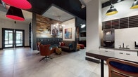 Located at 100 Union St in Elizabeth, NJ, this single-floor lobby space combines industrial and modern design elements to create a vibrant, welcoming communal environment. Large-format concrete-look tile flooring runs throughout, lending a clean, contemporary base to the space. Reclaimed wood accent walls add warmth and texture, contrasting beautifully with the dark-painted structural elements and exposed ceiling details overhead.

The lounge area is furnished with comfortable seating, including a tufted sofa and swivel chairs in rich earth tones, arranged beneath a striking geometric chandelier. Bold, colorful artwork — including a prominent pop-art-style piece — adorns the walls, adding personality and visual interest throughout. A bank of individual mailboxes is integrated seamlessly into the design along one wall.

A dedicated workstation area features a desk, computer, and wall-mounted television, while glass-partitioned spaces line one corridor, offering glimpses into adjacent rooms. The space also includes a ping-pong table beneath vibrant pendant lights in orange, pink, and red, alongside vending machines for added convenience. Large glass entry doors allow natural light to filter in, enhancing the open, airy feel of this well-appointed and thoughtfully designed communal lobby.