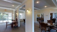 This single-floor space showcases a thoughtfully designed interior with classic architectural elements that lend it a refined, traditional character. Prominent white columns and wide doorway openings divide the layout into distinct functional areas while maintaining an open, connected feel throughout. Crown molding and coffered ceiling details add a sense of elegance and visual depth to each zone.

Wall-mounted sconce lighting provides warm, ambient illumination that complements the neutral, cream-toned walls, creating an inviting atmosphere across the space. The carpeted flooring runs consistently through the interior, contributing to a cohesive and comfortable environment.

To one side, a dining area is furnished with a wooden table and chairs, positioned near a window that allows natural light to filter in. On the opposite side, a seating area features upholstered and wooden chairs arranged for conversation or relaxation. In the background, a kitchen space is visible, equipped with cabinetry and a counter bar with seating, alongside what appears to be a living area with a sofa and additional furnishings. The overall ambiance is warm, classic, and well-organized.