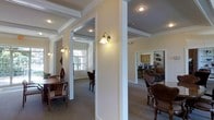 This single-floor space showcases a thoughtfully designed interior with classic architectural elements that lend it a refined, traditional character. Prominent white columns and wide doorway openings divide the layout into distinct functional areas while maintaining an open, connected feel throughout. Crown molding and coffered ceiling details add a sense of elegance and visual depth to each zone.

Wall-mounted sconce lighting provides warm, ambient illumination that complements the neutral, cream-toned walls, creating an inviting atmosphere across the space. The carpeted flooring runs consistently through the interior, contributing to a cohesive and comfortable environment.

To one side, a dining area is furnished with a wooden table and chairs, positioned near a window that allows natural light to filter in. On the opposite side, a seating area features upholstered and wooden chairs arranged for conversation or relaxation. In the background, a kitchen space is visible, equipped with cabinetry and a counter bar with seating, alongside what appears to be a living area with a sofa and additional furnishings. The overall ambiance is warm, classic, and well-organized.