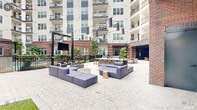 Located at 313 Liggett Street in Durham, NC, this amenity space offers a thoughtfully designed courtyard and indoor lounge experience set within a modern mixed-use building. The open-air courtyard is paved with distinctive hexagonal stone pavers and centers around a gated swimming pool, surrounded by lush greenery and manicured landscaping. Generous seating areas feature modular blue sectional sofas arranged for conversation and relaxation, while string lighting overhead adds ambiance for evening gatherings. An outdoor kitchen island with built-in grills, bar seating, and granite countertops provides an ideal setting for entertaining, complemented by long communal dining tables nearby. A turf lawn area accommodates lawn games such as cornhole, and the entire courtyard is framed by the building's striking blend of brick and contemporary paneling. Accessible via large glass sliding doors, the indoor lounge features exposed brick walls, a warm natural wood coffered ceiling, and statement chandeliers. Comfortable seating arrangements, a wall-mounted TV, and a wet bar make it a versatile space for social gatherings or casual lounging. Together, these indoor and outdoor spaces create a cohesive, resort-inspired amenity destination in the heart of Durham.