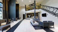 This impressive two-story space makes a bold architectural statement with its soaring double-height ceilings and dramatic open floor plan. The main level is anchored by a spacious lounge area furnished with contemporary light-gray sofas and sleek seating arrangements, creating an inviting yet refined atmosphere. A striking two-toned tile floor in white and deep charcoal adds a bold graphic element that defines the space.

At the rear, a full bar and kitchen area features a row of counter-height bar stools, pendant lighting in warm tones, and a decorative tile backsplash — ideal for entertaining or social gatherings. Dark cabinetry and muted finishes throughout lend a sophisticated, moody contrast to the light-colored surfaces.

The upper mezzanine level is accessed by a staircase and is enclosed by an ornate wrought-iron railing with geometric detailing, adding an elegant architectural focal point. Structural columns in dark tones punctuate the vertical expanse, reinforcing the contemporary design language. Recessed lighting and a wall-mounted flat-screen television complete the modern amenities. The cohesive palette of navy, gray, black, and white gives the entire space a polished, upscale ambiance suited for both relaxed lounging and more formal social functions.