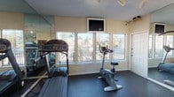 The Steeplechase fitness center is a well-equipped, single-floor exercise facility designed to support a range of workout routines. The space features a dark rubber flooring surface throughout, providing a durable and functional foundation suitable for both cardio and strength training activities. Large windows along one wall fill the room with natural light and offer open views of the surrounding community, including a fenced outdoor area and neighboring buildings. A wall-mounted flat-screen television is positioned above the windows, allowing residents to stay entertained during cardio sessions.

The cardio area is equipped with a treadmill, an upright stationary bike, and an elliptical machine. Floor-to-ceiling mirrors line multiple walls, visually expanding the space and providing a practical reference point during workouts. The strength training section features a multi-station cable and weight machine, offering a variety of resistance-based exercises. Track lighting along the ceiling supplements the natural light in the strength training area. A textured ceiling finish is consistent throughout the space. The facility is accessible via an exterior door that opens directly to the outdoor community grounds, providing convenient access for residents.