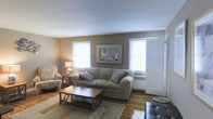 This single-floor residence offers a thoughtfully arranged living space designed for comfort and functionality. The living room serves as the heart of the home, showcasing warm hardwood flooring that adds a classic, inviting character to the space. Neutral wall tones in soft beige and taupe create a calming backdrop, complemented by a smooth, light-colored ceiling that enhances the sense of openness. Multiple windows flood the room with natural light, brightening the interior throughout the day. A baseboard heating unit ensures year-round comfort. The living area is well-proportioned, accommodating a full sofa suite, armchair, and a central coffee table atop a coordinating area rug, creating a cohesive and relaxed seating arrangement. A framed wall piece serves as a tasteful focal point above the main seating area. An entry door with a window panel is conveniently accessible from the living space, providing a seamless transition to the exterior. The overall ambiance is warm, understated, and practical, making it a welcoming environment suitable for everyday living.