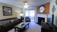 This single-floor living space features a well-appointed living room designed for comfort and functionality. The room is anchored by a classic white-mantled fireplace with a dark surround, adding a focal point of warmth and character to the space. An accent wall in a warm terracotta tone complements the neutral beige walls throughout, creating a layered and inviting color palette. A large round mirror with a dark wood frame adorns the accent wall, enhancing the sense of depth in the room.

Natural light fills the space through a sliding glass door dressed with sheer white vertical blinds, which opens to an outdoor area, providing a seamless connection to the exterior. The room is finished with plush carpeting underfoot, adding to the cozy atmosphere. A ceiling fan with a built-in light fixture is centrally positioned, ensuring both air circulation and ambient lighting. Furnishings include a dark upholstered sofa, a matching armchair, and a glass-top coffee table that reflects light throughout the room. Thoughtful decorative touches, including table lamps and subtle décor accents, complete this well-balanced living space.