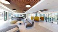 Located at the heart of Old Town Scottsdale, this expansive single-floor amenity space at Scottsdale on Main offers a sophisticated and thoughtfully designed environment spanning a generous footprint. The living and lounge area immediately impresses with its striking slatted wood ceiling panels, which frame a series of bold pendant light fixtures and create a warm architectural contrast against crisp white walls and herringbone-patterned light wood flooring. Floor-to-ceiling glass windows and sliding doors flood the interior with abundant natural light while offering direct views of the outdoor pool terrace and surrounding Scottsdale skyline. Flexible seating arrangements — including modular gray sofas, rounded ottomans, and bold yellow privacy chairs — provide distinct zones for relaxation and social interaction. The kitchen area features sleek, dark cabinetry with stone countertops and panoramic windows overlooking the pool deck, blending functionality with an open, airy aesthetic. A dramatic lobby staircase with glass railings, warm wood treads, and a terrazzo feature wall bearing the Scottsdale on Main logo adds a refined arrival experience. An artistic chandelier installation and full-height curtain glass walls in the atrium further elevate the sense of grandeur. Access to the Skyline Pool & Spa completes this premier amenity offering.