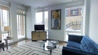 This single-floor living space offers a clean, modern aesthetic defined by an abundance of natural light. Large floor-to-ceiling windows line the walls, flooding the interior with daylight while framing views of the surrounding urban environment. The windows are fitted with roller shades, allowing for flexible light control throughout the day.

The living area showcases hardwood flooring paired with a plush, geometric-patterned area rug that anchors the seating arrangement. A sleek glass-top coffee table sits at the center, complemented by a tufted navy blue accent chair that adds a touch of sophistication. An ornate gold-toned media cabinet serves as both a functional and decorative focal point, supporting a flat-screen television.

A framed abstract artwork in earth tones — gold, grey, and black — adds a curated, gallery-like quality to the neutral white walls. The overall color palette is light and airy, with white walls and trim creating a crisp backdrop that highlights the carefully selected furnishings. The space conveys a refined urban sensibility, blending contemporary design with warm, inviting details.