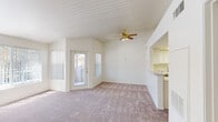 This single-floor residence offers a well-organized layout with a clean, neutral aesthetic throughout. The living area is the heart of the home, featuring vaulted ceilings that enhance the sense of openness, a gold-toned ceiling fan with integrated lighting, and soft carpeted flooring. A large front-facing window with vertical blinds and a glass-paneled entry door allow generous natural light to fill the space. The living area flows seamlessly into the kitchen via a pass-through counter, creating an open and connected feel. The kitchen is outfitted with white cabinetry, ample counter space, a full-size refrigerator, an electric range, and a sink — all set on durable wood-look flooring. The bedroom is a generously sized, carpeted room with vaulted ceilings and a window that brings in natural light. The full bathroom features a single-sink vanity with white cabinetry, a large wall mirror, a combined tub and shower with tiled surround, and wood-look flooring. A dedicated laundry area includes a washer and dryer with overhead wire shelving for added storage. Throughout the residence, the bright white walls, neutral tones, and mix of carpet and resilient flooring contribute to a clean and functional living environment.