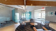 Located at 7061 Alvern St in Los Angeles, this single-floor commercial space delivers a compelling blend of industrial character and contemporary design. The architecture is defined by dramatic vaulted ceilings with exposed natural wood beams that span the open interior, creating an airy, loft-like atmosphere. Polished hardwood flooring runs throughout the main areas, complementing the warm tones of the structural beams overhead.

The central hub of the space is a generously sized open lounge area anchored by a circular area rug with a bold marbled pattern, furnished with mid-century-inspired blue upholstered chairs and round wooden tables — ideal for informal collaboration or casual meetings. Adjacent to this lounge, a glass-walled conference room with teal accent walls offers a visually striking yet acoustically separated space for formal gatherings.

Open workstation areas with individual desks line one side of the space, while decorative hexagonal acoustic panels in blue and neutral tones add both visual interest and sound absorption. Abstract artwork adorns the walls, reinforcing a creative, professional atmosphere.

Large glass doors and windows invite abundant natural light and provide views to an outdoor terrace. A kitchenette featuring grey cabinetry, white countertops, stainless steel appliances, and a teal accent wall rounds out the functional amenities of this well-appointed space.