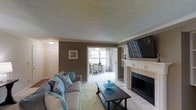 This single-floor space offers a welcoming and functional layout centered around a comfortable living room. The room features a classically designed fireplace with a white-painted mantle and tile surround, serving as a natural focal point. A flat-screen television is mounted above, making the space practical for everyday living. The walls are finished in a warm taupe tone, complemented by white crown molding and a textured ceiling that adds subtle architectural character. Soft, neutral carpeting flows throughout, contributing to the room's relaxed ambiance. Tasteful furnishings, including a plush sofa accented with teal and patterned throw pillows, add a touch of color and comfort. A dark wood coffee table anchors the seating area. An open doorway connects the living room to a dedicated home office or study, which benefits from a large window dressed with patterned drapes, allowing natural light to filter in. The workspace is equipped with a desk and chair setup. Near the entry, a white paneled door provides access to additional areas of the home, reinforcing the space's practical and well-organized flow.