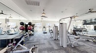 Located at 1392 E Bonds Ranch Rd in Fort Worth, Texas, this fitness center offers a generously sized, single-floor exercise space designed to support a wide range of workout routines. The room features stylish, textured carpet tiles and is illuminated by a combination of natural light from multiple windows and warm pendant lights, creating a bright and inviting atmosphere. Ceiling fans are distributed throughout, ensuring comfortable airflow during workouts. A full wall of floor-to-ceiling mirrors visually expands the space and includes a mounted flat-screen television, enhancing the workout experience. The facility is well-equipped with a comprehensive selection of cardio machines, including treadmills, ellipticals, a stair climber, and a stationary bike. Strength training is well represented with cable and resistance machines, a functional trainer, and a dedicated free weights area featuring a full dumbbell rack and medicine ball storage. The clean white walls and modern farmhouse-inspired lighting fixtures give the space a polished, contemporary aesthetic. Outdoor views through the windows add a sense of openness, while the thoughtful layout provides ample room to move freely between equipment stations.