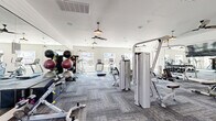 Located at 1392 E Bonds Ranch Rd in Fort Worth, Texas, this fitness center offers a generously sized, single-floor exercise space designed to support a wide range of workout routines. The room features stylish, textured carpet tiles and is illuminated by a combination of natural light from multiple windows and warm pendant lights, creating a bright and inviting atmosphere. Ceiling fans are distributed throughout, ensuring comfortable airflow during workouts. A full wall of floor-to-ceiling mirrors visually expands the space and includes a mounted flat-screen television, enhancing the workout experience. The facility is well-equipped with a comprehensive selection of cardio machines, including treadmills, ellipticals, a stair climber, and a stationary bike. Strength training is well represented with cable and resistance machines, a functional trainer, and a dedicated free weights area featuring a full dumbbell rack and medicine ball storage. The clean white walls and modern farmhouse-inspired lighting fixtures give the space a polished, contemporary aesthetic. Outdoor views through the windows add a sense of openness, while the thoughtful layout provides ample room to move freely between equipment stations.