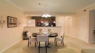 This single-floor residence offers a thoughtfully designed open-concept layout that seamlessly connects the dining area and kitchen, creating a sense of flow and spaciousness. The dining area is anchored by a stylish round dark-wood table surrounded by upholstered cream chairs, illuminated by an elegant multi-bulb chandelier that serves as a striking focal point overhead. Soft, neutral-toned carpeting and warm beige walls contribute to the inviting and cohesive ambiance throughout the space. The kitchen is positioned just beyond a half-wall counter that doubles as a breakfast bar, providing both visual separation and connectivity. Rich dark wood cabinetry lines the kitchen walls, complemented by pendant lighting that adds warmth to the cooking area. Black appliances, including a refrigerator and microwave, are neatly integrated into the cabinetry. A hallway visible in the background suggests additional private rooms, offering a sense of depth and functional layout. The overall design balances practicality with comfort, making it well-suited for both everyday living and entertaining.