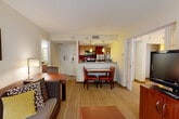 Located in Westborough, MA, this thoughtfully designed suite offers three bedrooms and three bathrooms across a single floor, making it an ideal space for extended stays or group accommodations. The open-concept living and dining area serves as the heart of the suite, featuring a comfortable sofa, armchair, and coffee table alongside a dedicated dining table with seating. Large windows dressed in sheer curtains allow natural light to fill the space, creating a warm and inviting atmosphere. The kitchen is efficiently laid out with stainless steel appliances, including a full-size refrigerator, range, and microwave. Rich red accent walls and warm pendant lighting add a contemporary touch, while a granite-topped breakfast bar connects the kitchen to the dining area seamlessly. Each bedroom is designed for comfort, featuring plush bedding, warm golden accent walls, rich wood furnishings including headboards and dressers, wall-mounted bedside lamps, and individual televisions. The primary bedroom includes a mirrored closet for added functionality. The bathrooms are finished with granite countertops, rich wood cabinetry, framed mirrors, and glass shelving, complemented by elegant patterned wallcoverings. Striped carpeting flows throughout, tying the cohesive, hotel-style aesthetic together.