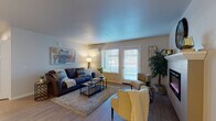 Located at 16548 NE Halsey St in Gresham, Oregon, this single-floor residence offers a well-designed layout with distinct living, dining, kitchen, and flexible bedroom spaces. The spacious living room is a central highlight, featuring wood-look flooring, a wall-mounted electric fireplace with a white mantel, a mini-split HVAC unit, and multiple windows dressed with white blinds that allow generous natural light to fill the room. The kitchen is appointed with rich dark wood cabinetry, light countertops, stainless steel appliances including a refrigerator, range, and over-the-range microwave, and durable hard-surface flooring. Adjacent to the kitchen is a dedicated dining area with wood-look floors, offering an intimate setting for meals. The bathroom features light wood-look flooring, dark wood vanity cabinetry, a single-sink vanity with a large mirror, and a clean, open layout. Carpeted rooms provide versatile spaces suitable for bedrooms or a home office, each with neutral-toned walls and natural light from windows with blinds. The overall ambiance is modern and comfortable, with a cohesive neutral color palette, practical finishes, and a functional open-concept flow throughout the single-level floor plan.