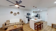 This single-floor space showcases a thoughtfully designed open-concept layout that seamlessly integrates the living and kitchen areas. Light wood-tone flooring runs continuously throughout, enhancing the bright, airy atmosphere created by white walls and recessed lighting. A ceiling fan anchors the living area, which features an electric fireplace with a wood mantel, adding warmth and character to the space.

The kitchen is a standout feature, outfitted with crisp white shaker cabinets, black hardware, and quartz countertops. A farmhouse-style apron sink sits beneath a window that allows natural light to fill the space. Stainless steel appliances, including a refrigerator, range, and microwave, complement the contemporary design. A central island with a white quartz top and bar seating, illuminated by decorative cage pendant lights, provides an ideal casual dining and prep area.

The bedroom is a comfortable retreat with natural light from slatted blinds, a ceiling fan, and warm wood flooring. The bathroom features hexagonal tile flooring, a quartz-topped white vanity, black fixtures, and a tub-shower combination with a sliding glass door, maintaining the clean, modern aesthetic found throughout the entire space.