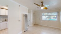 This single-floor space offers a clean, bright aesthetic throughout, defined by light-toned wood flooring, crisp white walls, and elegant crown molding that lend a polished, cohesive feel to every room. The main living area is open and airy, bathed in natural light from a window overlooking an outdoor area with a visible railing. A contemporary multi-blade ceiling fan with a brass-toned fixture serves as a stylish focal point while providing practical ventilation. A wall-mounted heating unit ensures year-round comfort. A closet with louvered doors adds convenient storage directly within the living space. The kitchen is a standout feature, showcasing white shaker-style cabinetry, granite countertops with a matching full stone backsplash, a stainless steel sink, and a gas range — offering both functionality and refined aesthetics. The bathroom is finished in a clean, neutral palette with white tile surrounding a full bathtub and shower combo, a window for natural light and ventilation, and brushed nickel fixtures throughout. Overall, the space presents a well-maintained, move-in-ready environment with thoughtful finishes that balance practicality and modern style.