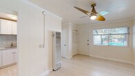This single-floor space offers a clean, bright aesthetic throughout, defined by light-toned wood flooring, crisp white walls, and elegant crown molding that lend a polished, cohesive feel to every room. The main living area is open and airy, bathed in natural light from a window overlooking an outdoor area with a visible railing. A contemporary multi-blade ceiling fan with a brass-toned fixture serves as a stylish focal point while providing practical ventilation. A wall-mounted heating unit ensures year-round comfort. A closet with louvered doors adds convenient storage directly within the living space. The kitchen is a standout feature, showcasing white shaker-style cabinetry, granite countertops with a matching full stone backsplash, a stainless steel sink, and a gas range — offering both functionality and refined aesthetics. The bathroom is finished in a clean, neutral palette with white tile surrounding a full bathtub and shower combo, a window for natural light and ventilation, and brushed nickel fixtures throughout. Overall, the space presents a well-maintained, move-in-ready environment with thoughtful finishes that balance practicality and modern style.