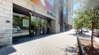 Located within The Aspen, a contemporary residential building with a striking exterior of stone cladding and floor-to-ceiling glass, this single-floor unit offers a thoughtfully designed interior with a clean, modern aesthetic. The building's street-level entrance is welcoming, featuring curated landscaping with bold planters and a well-maintained paved walkway.

Inside the unit, warm hardwood-style flooring runs throughout, complementing the cool gray-toned walls and crisp white trim. One of the most distinctive design elements is the frosted glass sliding barn door, which adds a stylish, semi-transparent partition between spaces while allowing natural light to filter through — a detail that balances privacy with an open feel.

The unit includes an in-unit stacked washer and dryer, a practical and convenient amenity tucked neatly into a dedicated space. The full bathroom features a bathtub and shower combination, a toilet, and a towel bar, all finished in a simple, clean style consistent with the rest of the unit.

Natural light plays a notable role in the ambiance, with sunlight casting bright, angled patterns across the walls and floors. The overall layout is functional and well-proportioned, with multiple doorways connecting the spaces in a straightforward, easy-to-navigate flow. The unit presents a polished, move-in-ready environment suited to modern urban living.