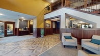 This welcoming lobby is defined by its striking architectural design and refined finishes. At the heart of the space, an eye-catching geometric-patterned hardwood floor creates a bold visual statement, transitioning seamlessly into carpeted areas throughout. Soaring double-height ceilings with bold golden-yellow accent walls draw the eye upward, where a mezzanine-level overlook features dark wood railings and a staircase with wrought iron balusters, adding vertical drama to the open atrium.

Rich dark wood wainscoting lines the walls throughout, lending warmth and a polished, traditional character to the space. A front desk reception area is centrally positioned, flanked by elevator access and a mailroom alcove fitted with individual mail compartments and a coffee station, enhancing the space's functional amenities.

Adjacent seating areas offer comfortable, stylishly furnished nooks, including an intimate booth-style lounge near large windows that flood the area with natural light and views of mature outdoor greenery. A secondary corridor features track lighting, decorative wall mirrors, and a display console, guiding visitors deeper into the building. Throughout, the careful layering of textures, from wood paneling to patterned carpet and tile, creates a cohesive and inviting atmosphere befitting a well-appointed communal building entry.