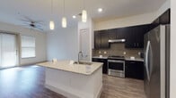 This well-designed single-floor residence offers a thoughtful layout that balances functionality and modern style. The open-concept kitchen serves as the heart of the space, showcasing rich dark cabinetry that extends to the ceiling, a neutral tile backsplash, and light granite countertops. A central island with an integrated sink and elegant pendant lighting above provides an ideal space for meal prep or casual dining. Stainless steel appliances, including a range, refrigerator, and microwave, complete this polished kitchen. Wood-look flooring runs throughout the main living areas, lending warmth and continuity to the space.

The living area connects seamlessly to the kitchen and features built-in shelving, offering both storage and display opportunities. A ceiling fan overhead adds comfort to the open layout.

The bedroom is bright and inviting, filled with natural light streaming through large windows fitted with horizontal blinds. Plush carpet underfoot adds a cozy touch, and the room connects directly to a generous walk-in closet with wrap-around hanging rods and a carpeted floor.

The bathroom features a dark-stained vanity with a granite countertop, a large mirror with contemporary lighting, and light-toned tile flooring. Accessible grab bars are thoughtfully incorporated, reflecting an inclusive design approach throughout this well-appointed space.