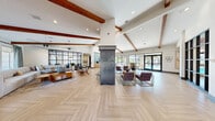 Located at 9270 Eagle Ranch Rd NW in Albuquerque, New Mexico, this single-floor communal living space is a beautifully designed gathering area that blends contemporary style with warm, inviting details. The open-plan layout is anchored by stunning herringbone-patterned light wood flooring that flows seamlessly throughout the entire space, creating a cohesive and polished aesthetic.

Soaring vaulted ceilings with exposed rustic wood beams lend architectural character and a sense of grandeur to the interior. A dramatic freestanding fireplace clad in dark metal serves as a bold focal point, flanked by curated seating and topped with a large-scale piece of artwork. Statement pendant lighting, including sculptural vertigo-style fixtures, adds visual interest overhead.

A spacious lounge area features a generous sectional sofa and eclectic accent chairs arranged for comfortable socializing. Steel-framed glass partition walls define a private meeting or office area while maintaining an open, airy feel. A bar or counter area with pendant lighting and modern cabinetry offers additional functionality.

Natural light filters in through multiple windows and glass entry doors, highlighting the crisp white walls adorned with decorative wall art and sculptural accents. The overall ambiance is modern yet approachable, combining sophisticated design elements with comfortable, community-oriented spaces.