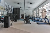 Located at 471–483 N McDonough St in Decatur, Georgia, this single-level fitness center is designed to support a comprehensive range of workout routines. The main exercise floor is expansive and bathed in natural light from floor-to-ceiling steel-framed windows that offer views of the surrounding streetscape. Soaring ceilings with ceiling fans enhance airflow and contribute to the open, airy atmosphere of the space. The main floor is outfitted with a variety of strength training machines and cardio equipment, including treadmills and cable machines, arranged across a combination of carpet and hard flooring surfaces. Large mirrors visually expand the space and reflect the abundant natural light throughout. Bold athletic murals and motivational graphics are displayed prominently on the walls, reinforcing the energetic character of the facility. A dedicated spin studio, accessible through glass-framed doors, features stationary cycling bikes, wall-mounted accessories, kettlebell storage, and mirrored walls with a ballet barre, creating a focused environment for group or individual cycling sessions. A separate free weights area with a tufted blue carpet floor, a weight bench, and a dumbbell rack provides a quieter zone for strength-focused training. Together, these distinct zones create a dynamic and functional fitness environment.