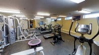 This fitness center is a purpose-built exercise facility designed to support a well-rounded workout routine. The space features durable rubber flooring throughout, providing both safety and comfort during exercise. A large wall-mounted mirror spans one side of the room, enhancing the sense of space and allowing users to monitor their form during workouts.

The cardio equipment selection includes a treadmill, an elliptical trainer, a stair climber, and an upright stationary bike, offering a range of options for cardiovascular training. For strength and resistance work, the room is equipped with a multi-functional weight machine and a bench press setup, accommodating various muscle groups and training styles. A mini trampoline rounds out the equipment, providing a fun, low-impact cardio alternative.

The room is illuminated by recessed fluorescent lighting set into a textured ceiling, ensuring a bright and well-lit environment. Small windows positioned near the ceiling allow for a degree of natural light. A vending machine is conveniently located in the corner, providing easy access to refreshments. The walls are finished in a neutral tone, contributing to a clean and functional atmosphere throughout the space.