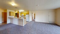 Located on Hidden Hills Drive in Kalamazoo, Michigan, this single-floor unit offers a practical, well-organized layout designed for comfortable everyday living. The open-concept design connects the kitchen and living area seamlessly, with a peninsula counter serving as both a workspace and casual dining surface. The kitchen is equipped with light oak cabinetry, white appliances including a refrigerator, electric range, and built-in microwave, complemented by a double stainless steel sink and tile-patterned flooring. A dedicated laundry alcove within the kitchen area accommodates a full-size washer and dryer. The spacious living area features plush carpeting and vertical blinds that allow natural light to filter through. Bedrooms are carpeted and include windows with blinds, offering natural light; one bedroom features a built-in wall-mounted desk shelf, while another includes a standard closet with a wood-panel door. The bathroom is appointed with a single-sink vanity featuring warm wood cabinetry, a large mirror with overhead lighting, and a full-size tub and shower combination. Textured ceilings and neutral wall tones throughout lend the space a clean, consistent aesthetic.