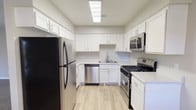 This single-floor space showcases a well-appointed kitchen designed with a clean, contemporary aesthetic. White shaker-style cabinetry lines the walls from base to upper level, providing generous storage throughout. The cabinetry is complemented by sleek bar-pull hardware, contributing to a polished, modern look. Stainless steel appliances include a gas range, built-in microwave, dishwasher, and a full-size refrigerator, offering a fully equipped cooking environment. The layout follows a functional U-shaped configuration, maximizing counter space and workflow efficiency. A stainless steel sink is positioned beneath upper cabinets along the back wall, keeping the workspace organized and practical. Light wood-look flooring runs throughout, adding warmth and contrast against the white cabinetry and neutral grey walls. Overhead, a large flush skylight-style ceiling fixture provides bright, even illumination across the entire kitchen. The overall ambiance is fresh and airy, with a cohesive color palette of white, grey, and warm wood tones that creates an inviting and functional space well-suited for everyday cooking and entertaining.
