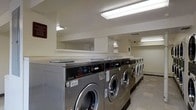 This commercial laundry facility is designed with functionality and efficiency in mind. The single-floor space is outfitted with a long row of stainless steel front-load commercial washing machines lining one wall, complemented by a matching set of dryers, providing ample capacity for high-volume laundry needs. The machines are modern SpeedQueen commercial-grade units, built for durability and consistent performance. Overhead fluorescent lighting illuminates the entire space evenly, ensuring a bright and practical environment. The walls are finished in a clean white, contributing to a crisp, hygienic atmosphere. A continuous countertop surface runs along the top of the machines, offering a convenient space for sorting and folding laundry. The flooring is durable and easy to maintain, suited to the demands of a busy facility. A security camera is visible, adding an element of safety and oversight to the space. The overall layout is straightforward and uncluttered, maximizing usable floor space while keeping all equipment easily accessible. This facility is well-suited for residential building tenants, hospitality operations, or any setting requiring reliable, large-scale laundry services.