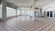 The Kirk Hotel's single-floor space presents a compelling combination of historic architectural details and open, adaptable layout. The most striking feature is the expansive intricate mosaic tile flooring, displaying an ornate geometric and floral pattern in muted earth tones—terracotta, cream, and taupe—that extends across the main gathering area, serving as a strong visual centerpiece. A curved, sculptural white plaster partition wall divides portions of the space, adding an organic, Art Deco-influenced architectural element that contrasts with the otherwise rectilinear structure.

Large glass block walls run along one side, diffusing abundant natural light throughout the interior while maintaining privacy, contributing to the bright, airy atmosphere. Structural columns are positioned throughout, and pendant globe light fixtures hang from the ceiling, reinforcing the period aesthetic. The flooring transitions from mosaic tile to polished concrete in a secondary area, suggesting distinct functional zones within the single floor.

A kitchen area is fitted with warm oak cabinetry featuring glass-front upper cabinets, black tile countertops with decorative border tiling, and a substantial island with ornate black metalwork detailing. Additional windows with transoms allow supplementary natural light. Overall, the space carries a layered, historically rich ambiance suited for a variety of uses.