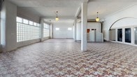 The Kirk Hotel's single-floor space presents a compelling combination of historic architectural details and open, adaptable layout. The most striking feature is the expansive intricate mosaic tile flooring, displaying an ornate geometric and floral pattern in muted earth tones—terracotta, cream, and taupe—that extends across the main gathering area, serving as a strong visual centerpiece. A curved, sculptural white plaster partition wall divides portions of the space, adding an organic, Art Deco-influenced architectural element that contrasts with the otherwise rectilinear structure.

Large glass block walls run along one side, diffusing abundant natural light throughout the interior while maintaining privacy, contributing to the bright, airy atmosphere. Structural columns are positioned throughout, and pendant globe light fixtures hang from the ceiling, reinforcing the period aesthetic. The flooring transitions from mosaic tile to polished concrete in a secondary area, suggesting distinct functional zones within the single floor.

A kitchen area is fitted with warm oak cabinetry featuring glass-front upper cabinets, black tile countertops with decorative border tiling, and a substantial island with ornate black metalwork detailing. Additional windows with transoms allow supplementary natural light. Overall, the space carries a layered, historically rich ambiance suited for a variety of uses.