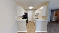 This single-floor 2-bedroom, 2-bathroom residence offers a clean, modern aesthetic throughout. The kitchen is a standout feature, designed in a galley-style layout that maximizes efficiency and storage. Crisp white cabinetry lines both sides of the kitchen, complemented by light quartz-style countertops that extend into a convenient breakfast bar, creating a natural transition into the living area. Black appliances, including a gas range, microwave, and refrigerator, provide a striking contrast against the bright white finishes. A stainless steel sink with a modern faucet sits beneath upper cabinetry on the opposite wall, offering ample workspace. Warm wood-look flooring runs throughout, adding texture and continuity to the space. The kitchen is well-lit with flush-mounted ceiling lighting and under-cabinet illumination. Neutral gray walls keep the palette versatile and inviting. A door at the end of the kitchen leads to additional areas of the unit, suggesting a thoughtful, functional floor plan. The overall ambiance is fresh, clean, and move-in ready, reflecting a contemporary residential design.